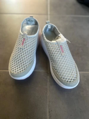 Grå mesh slip-on sneakers - Lätta och luftiga slip-on sneakers i grå mesh med vit sula. Skorna har ett sportigt och avslappnat utseende med textdetalj på ovandelen. Perfekta för dig som gillar bekväma och stilrena sneakers med modern känsla.