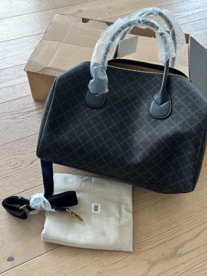 Lullu bag Mörkbrun weekendbag från By Malene Birger - Snygg weekendbag i mörkbrunt med diskret rutmönster och svarta detaljer. Väskan har guldfärgade metalldetaljer, avtagbar axelrem och två handtag. Tillverkad i tåligt material och levereras med dustbag.