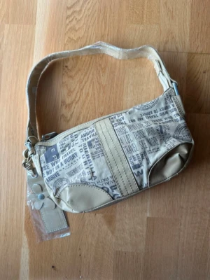 Beige axelväska med textmönster - Cool beige axelväska med tryckt textmönster i grått och vitt. Väskan har en bred axelrem och detaljer i konstläder. Praktisk storlek för det viktigaste och unik design med res- och stämpelmotiv. Perfekt för dig som gillar streetstyle och vill sticka ut.