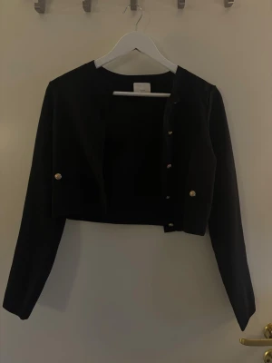 Svart kort kofta från H&M - Stilren svart kort kofta från H&M med långa ärmar och dekorativa guldfärgade knappar framtill. Perfekt att slänga över en topp för en snygg lager-på-lager-look. Enkel och klassisk design som passar till mycket.Hör av dig om du har några frågor! 💗🫶🏼