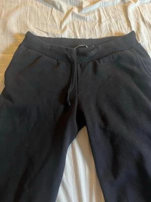 Svarta mjukisbyxor med dragsko - Säljer ett par svarta mjukisbyxor med resår i midjan och dragsko för skön passform. Byxorna har sidofickor och är gjorda i mjukt bomullsmaterial. Perfekta för chill dagar hemma eller när du vill vara bekväm.
