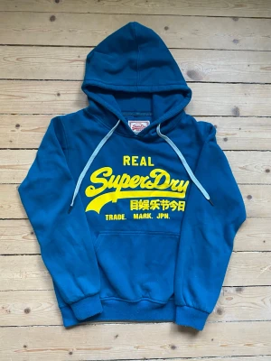 Superdry Hoodie  - Säljer denna blå/turkos Superdry Hoodie med ett gult tryck. Passar storlek M. Skick 8/10. Hör av dig vid minsta fråga. 