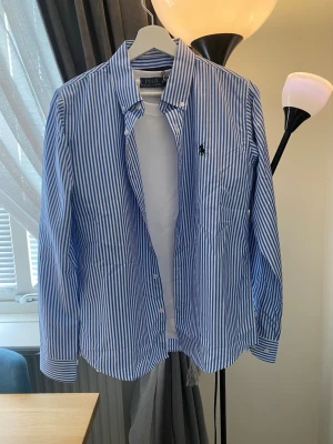 Blårandig skjorta från Ralph Lauren - Klassisk blårandig skjorta från Ralph Lauren med vit botten och blå vertikala ränder. Skjortan har knappar framtill, lång ärm och den ikoniska broderade loggan på bröstet. Tillverkad i bomull och har en tidlös, normal passform.
