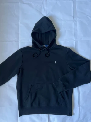 Svart hoodie från Polo Ralph Lauren - Klassisk svart hoodie från Polo Ralph Lauren med huva och dragsko. Har en broderad vit logga på bröstet och en stor magficka framtill. Tillverkad i mjukt bomullsmaterial och har ribbade muddar vid ärmslut och nederkant.
