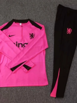 Rosa Nike träningsset med logga - Snyggt träningsset från Nike med långärmad rosa tröja och svarta byxor med rosa detaljer. Tröjan har halv dragkedja, svarta axelpartier och klubbmärke på bröstet. Byxorna är svarta med rosa logga och inslag längs benen. Perfekt för fotbollsträning eller gym.