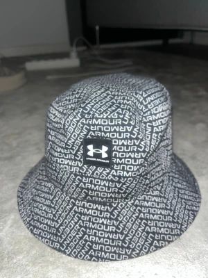Svart bucket hat från Under Armour - Cool svart bucket hat från Under Armour med vitt logomönster över hela hatten och en tydlig logopatch framtill. Hatten är tillverkad i bomull och polyester, perfekt för att lyfta din streetstyle. Snygg och trendig design som sticker ut.