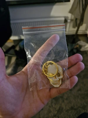 Nyckelring med dödskalle i guld - Säljer en cool nyckelring i metall med dödskallemotiv. Nyckelringen är guldfärgad med vita detaljer och har en rund ring för att fästa nycklar eller accessoarer. Perfekt för dig som gillar edgy detaljer till din stil.