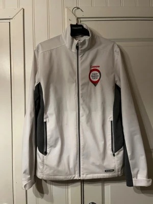 Vit golf jacka  - Vit softshelljacka från Canon med broderad logga och texten 'Canon Business Center' på bröstet. Jackan har svarta detaljer på sidorna, dragkedja framtill och två fickor med dragkedja. Perfekt för dig som gillar stilren och sportig look.