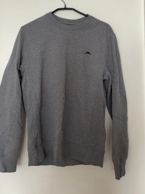 Grå sweatshirt från J.Lindeberg - Klassisk grå sweatshirt från J.Lindeberg med rund halsringning och diskret svart JL-logga broderad på bröstet. Tröjan har långa ärmar och ribbade muddar vid ärmslut och nederkant. Perfekt för en avslappnad och stilren look.