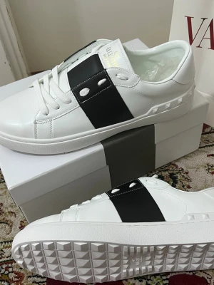 Valentino Garavani skor  - Snygga vita sneakers från Valentino Garavani med bred svart läderdetalj och nitar på sidan. Skorna har vita snören, låg profil och coola detaljer på sulan. Perfekta för dig som vill ha en stilren men ändå unik look.