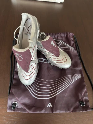 Adidas Predator Elite Bellingham  - Säljer ett par Adidas Predator Elite Bellingham. Användna 2-3gånger. fotbollsskor i silver med vinröda detaljer och vita inslag. Skorna har klassisk snörning, gummidetaljer för bättre bollkontroll och FG-dobbar för spel på naturgräs. Medföljer matchande vinröd påse med Predator-logga.