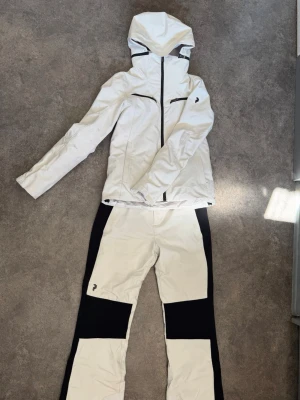 Peak performance skidställ - Säljer ett snyggt och stilrent skidställ från Peak Performance i offwhite/vit färg med svarta detaljer. Setet består av jacka i storlek S och byxor i storlek M. Det är använt under en säsong men är fortfarande i bra skick och fullt fungerande.  Jackan är utrustad med Peak Performances HIPE Core+. Det finns vissa smutsmärken och fläckar, främst på det vita tyget, vilket syns på bilderna. Kan tänka mig att det går bort i tvätt!