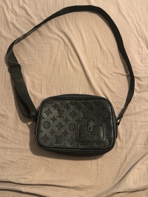 Svart axelväska från H&M - Snygg svart axelväska från H&M med klassiskt monogrammönster över hela väskan. Väskan har justerbar axelrem och en praktisk ficka framtill med knapp. Tillverkad i präglat läder och har en stilren, rektangulär form.