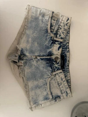 Blåa lågmidjade denimshorts med fransig kant - Säljer ett par ljusblå denimshorts från Tally weijl med fransig kant och klassisk femficksdesign. Shortsen har låg midja och en tvättad, nästan blekt look som ger en cool vintagekänsla. Perfekta för varma dagar och enkel styling.
