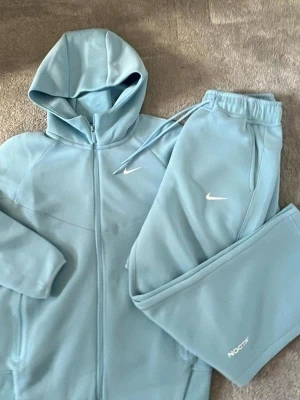 Ljusblå Nike NOCTA mjukisdress - Säljer en ljusblå mjukisdress från Nike NOCTA-kollektionen. Setet består av en hoodie med dragkedja och matchande byxor med dragsko i midjan. Båda delarna har vita Nike-loggor och byxan har NOCTA-tryck. Perfekt för chill dagar hemma.