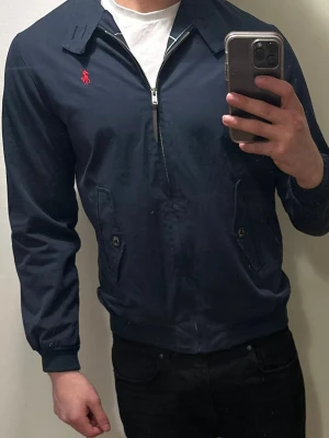 Marinblå bomberjacka Polo Ralph Lauren - Snygg marinblå bomberjacka från Polo Ralph Lauren med röd broderad logga på bröstet. Jackan har dragkedja, två fickor med knapp och ribbade muddar. Insidan är fodrad med rutigt tyg för extra stilpoäng. Perfekt för dig som vill ha en klassisk och clean look.