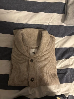 Beige kofta med sjalkrage från Uniqlo - Säljer en beige ribbstickad kofta från We Fashion med sjalkrage och bruna knappar. Koftan har två fickor framtill och är tillverkad i ett mjukt, tjockare material som passar perfekt för kyligare dagar. Enkel och stilren design som är lätt att matcha.