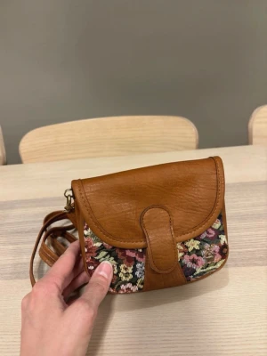Brun axelväska med blommigt tyg - Säljer en liten axelväska i brunt läder med detaljer i blommigt tyg. Väskan har en rundad form, justerbar axelrem och ett snyggt guldfärgat spänne framtill. Perfekt för dig som gillar retrostil och unika accessoarer.               Mått  Bredd 15cm Höjd 13cm Djup 5cm  Ett ytterfack med dragkedja baksida väskan och inuti är det ett fack samt ett mindre fack.