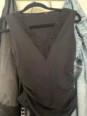 Svart body med spetsdetalj - Snygg svart body med djup V-ringning och spetskant längs urringningen. Ärmlös modell i mjukt stretchigt material som sitter tight och bekvämt. Perfekt att styla med jeans eller kjol för en trendig look.