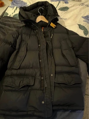 Svart dunjacka från Parajumpers - Snygg svart dunjacka från Parajumpers med huva och stora fickor framtill. Jackan har en gul detalj vid halsen och grått innerfoder. Perfekt för kalla dagar och har en klassisk, puffig design med dragkedja och tryckknappar.