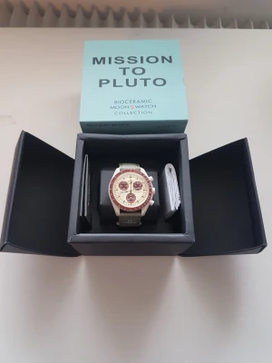 Omega x Swatch Mission to Pluto klocka - Säljer en Omega x Swatch Mission to Pluto från Bioceramic MoonSwatch Collection. Klockan har en beige urtavla med vinröda detaljer och grått armband. Snygg och unik design med kronograffunktion och tydliga index. Perfekt för dig som gillar statement-accessoarer.