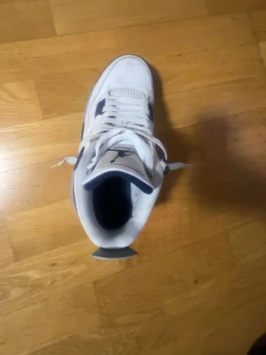 Vita Air Jordan 4 Retro sneakers - Säljer ett par vita Air Jordan 4 Retro sneakers med blå detaljer och klassisk Jumpman-logga på plösen. Skorna har meshpaneler på sidorna, robust sula och högt skaft. Perfekta för dig som gillar streetwear och ikoniska modeller.