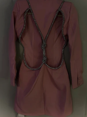 Rosa blazerklänning med öppen rygg och diamantdetaljer - Bella Barnett - Backless Diamond Blazer Dress  Trendig rosa kortklänning med lång ärm och kavajkrage är ett riktigt statement-plagg. Den öppna ryggen och sidutskärningarna med diamant detaljer ger ett exklusivt och modernt uttryck. Perfekt för fest, kvällsevent och andra speciella tillfällen. Kan bäras som miniklänning eller stylas med byxor för en trendig look.