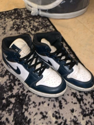 Nike Air Jordan 1 Mid Navy/White - Snygga Nike Air Jordan 1 Mid sneakers i marinblått och vitt med svarta detaljer och klassisk swoosh. Skorna har perforerad tå, svart snörning och ikonisk Jordan-logga på plösen. Perfekta för dig som gillar streetwear och sportig stil. Dåligt kvalite på skorna men värt priset.