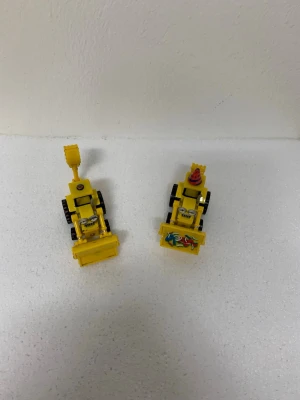 Leksaksfordon - Två coola gula leksakstraktorer med svarta hjul och detaljer i silver. Ena traktorn har ett färgglatt motiv med hästar på skopan och en röd/blå kon på taket. Båda har rörliga delar och roliga ansikten framtill. Perfekt för dig som gillar maskiner och samlar på unika leksaker.