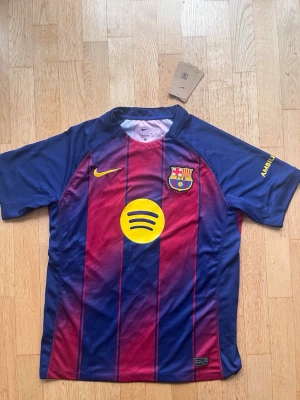 FC Barcelona matchtröja Nike blå/röd - Säljer en officiell FC Barcelona matchtröja från Nike i blått och rött med gula detaljer. Tröjan har korta ärmar, klubbmärke, Nike-logga och Spotify som sponsor framtill. På ryggen står det Lamine Yamal och nummer 10 i gult. Perfekt för dig som älskar fotboll!
