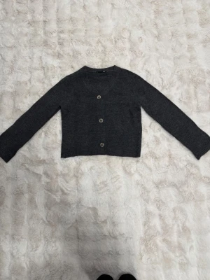 Mörkgrå kofta från 157, S - Mysig mörkgrå kofta från 157 med rund halsringning och tre knappar framtill. Kortare modell med långa ärmar, perfekt att slänga över en topp. Tillverkad i mjukt stickat material som känns skönt mot huden. Priset kan diskuteras 🩷