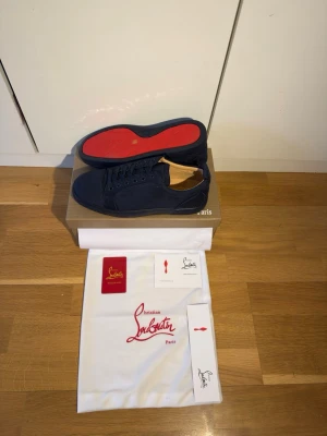 Marinblå Christian Louboutin sneakers - Snygga marinblå sneakers från Christian Louboutin med klassisk röd sula och diskret logga. Skorna är låga och har en stilren design i mocka med matchande snörning. Perfekta för dig som vill ha något exklusivt och trendigt i garderoben.