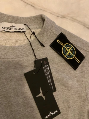 Grå sweatshirt från Stone Island - Snygg grå sweatshirt från Stone Island med klassisk patch på vänster ärm. Tröjan har rund halsringning, ribbade muddar och är tillverkad i mjuk bomull. Perfekt för en avslappnad och stilren look.