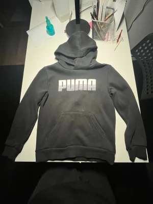 Svart hoodie från Puma med logga - Snygg svart hoodie från Puma med stor vit logga på bröstet. Klassisk känguruficka framtill och huva. Tillverkad i mjukt material som passar perfekt för en avslappnad stil. Perfekt för dig som gillar streetwear och sportiga vibes.