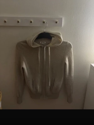 Beige hoodie med dragkedja Soft Goat - Mysig beige hoodie från Soft Goat i mjuk och lyxig kashmir. Hoodien har dragkedja framtill, två fickor och justerbar huva med snören. Perfekt för en avslappnad och stilren look.