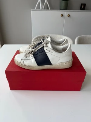 Valentino opens - Snygga vita sneakers från Valentino med bred mörkblå detalj på sidan och vita skosnören. Skorna har en klassisk låg siluett och är tillverkade i läder med gummisula. Perfekta för en stilren och trendig look. Med några defekter där av priset.Kan absolut gå ner i pris vid snabb affär.
