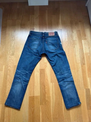 Levi's blå raka jeans - Klassiska blå jeans från Levi's med raka ben och femficksmodell. Jeansen har slitningar och whiskers på baksidan av knäna samt en brun läderpatch med logga bak i midjan. Tillverkade i denim med tydliga kontrastsömmar.Midjan är 39 cm (dubbla ca:78cm) innerbenslängd:69cm ytterbenslängd:96cm