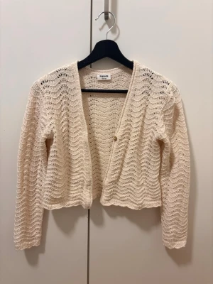 Beige virkad kofta från Kappahl - Söt beige kofta från Kappahl med vågigt virkat mönster och en knapp framtill. Koftan är kortare i modellen, har långa ärmar och är tillverkad i ett lätt, luftigt material som passar perfekt till lager på lager.