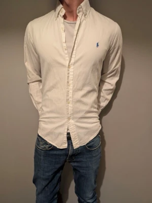 Vit skjorta från Ralph Lauren - Klassisk vit skjorta från Ralph Lauren med custom fit och blå logga broderad på bröstet. Skjortan har lång ärm, knappar framtill och en tidlös design som passar till jeans eller chinos. Tillverkad i bomull för en skön känsla.