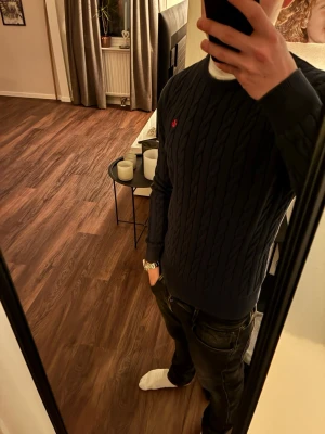 Mörkblå kabelstickad tröja Morris  - Snygg mörkblå kabelstickad tröja från Morris med rund halsringning och diskret röd logga broderad på bröstet. Tröjan har långa ärmar och ribbade muddar vid ärmslut och nederkant. Perfekt för en stilren och klassisk look.