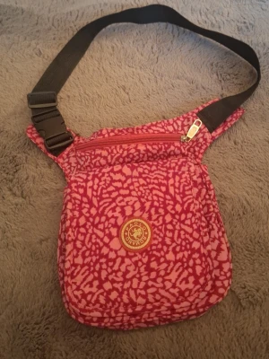 Rosa axelväska från Bomani - Axelväska i tyg från Bomani med rosa och ljusrosa mönster. Väskan har justerbar svart axelrem, dragkedja upptill och ett lock med kardborrestängning. Perfekt för att bära det viktigaste och ger en färgklick till din outfit.