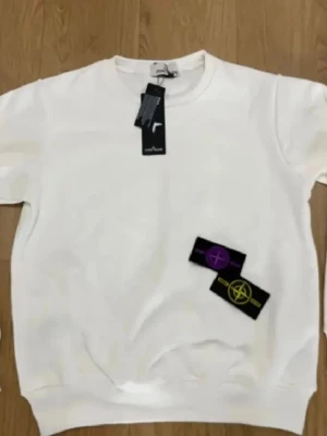 Vit sweatshirt från Stone Island - Snygg vit sweatshirt från Stone Island med klassisk rund hals och ribbade muddar. Medföljer två avtagbara logopatchar i svart med lila och gul brodyr. Perfekt för dig som vill ha en clean och stilren look med exklusiv känsla.