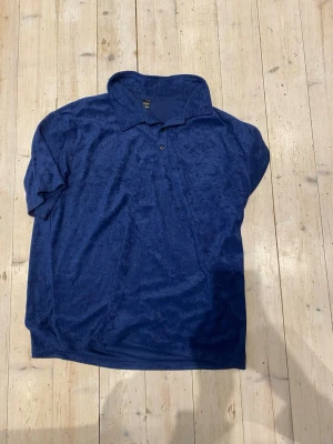 Blå polotopp i sammet - Mjuk och lyxig mörkblå polotopp i sammet med klassisk krage och korta ärmar. Toppen har knappar framtill och en avslappnad passform som ger en chill vibe. Perfekt för dig som vill sticka ut med en unik textur och djup färg.