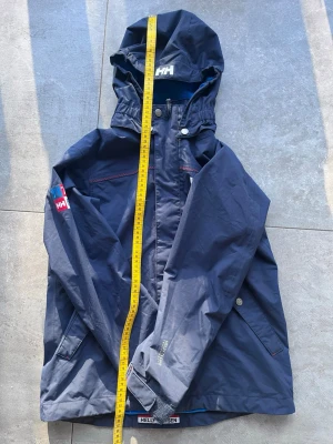 Helly Hansen Helly Tech® Jacka – Stl 164/14 år – Mörkblå – Nyskick - Högkvalitativ skaljacka från Helly Hansen i mörkblått med röda detaljer. Jackan är utrustad med "Helly Tech® Performance"-teknologi vilket innebär att den är helt vind- och vattentät samtidigt som den andas. Perfekt som vår/höstjacka eller som ett yttre lager vid friluftsaktiviteter. Avtagbar huva och reglerbara ärmslut. • Storlek: 164 / 14 år (Regular fit). • Mått: Se bilder (Längd ca 70 cm). • Skick: Nyskick, inga tecken på användning.