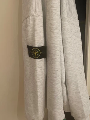 Ljusgrå hoodie från Stone Island - Säljer en ljusgrå hoodie från Stone Island med dragkedja framtill och klassisk logopatch på ärmen. Hoodien har justerbar huva med snören och är tillverkad i mjukt bomullsmaterial. Perfekt för dig som gillar streetwear och vill ha något stilrent.