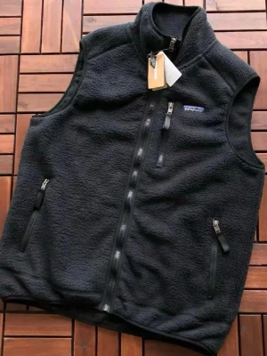 Svart fleeceväst från Patagonia - Mysig svart fleeceväst från Patagonia med hög krage och hel dragkedja framtill. Västen har tre praktiska fickor med dragkedja, en på bröstet och två på sidorna. Tillverkad i mjukt och värmande fleecematerial, perfekt för lager-på-lager-stil.