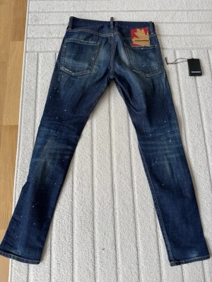 Mörkblå jeans från Dsquared2 - Säljer ett par mörkblå jeans från Dsquared2 med slitningar och färgstänk för en cool, trendig look. Klassisk femficksmodell med raka ben och kontrastsömmar. Jeansen har en röd Dsquared2-tag vid fickan och är tillverkade i denim. Kvitto finns!!