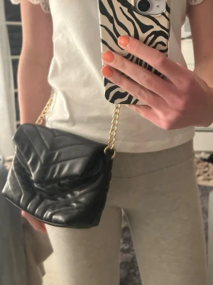 Svart handväska med kedja - Trendig svart quiltad handväska i mjukt material med guldfärgad kedjerem. Väskan har ett lock med V-format mönster och stängs med magnetknapp. Perfekt storlek för det viktigaste och ger en cool touch till din outfit.
