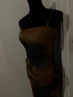 Mönstrad omlottklänning med smala band - Cool klänning med smala axelband och omlottdetalj i midjan. Färgerna går i mörkgrön, brun och beige med ett abstrakt mönster. Klänningen har en kroppsnära passform och är gjord i ett mjukt, stretchigt material.