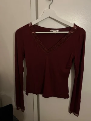 Vinröd långärmad topp med spets - Vinröd långärmad topp med v-ringning och spetsdetaljer längs halsringningen och ärmsluten. Tunn och mjuk, perfekt för lager på lager eller som den är. Enkel men elegant design som passar till mycket.